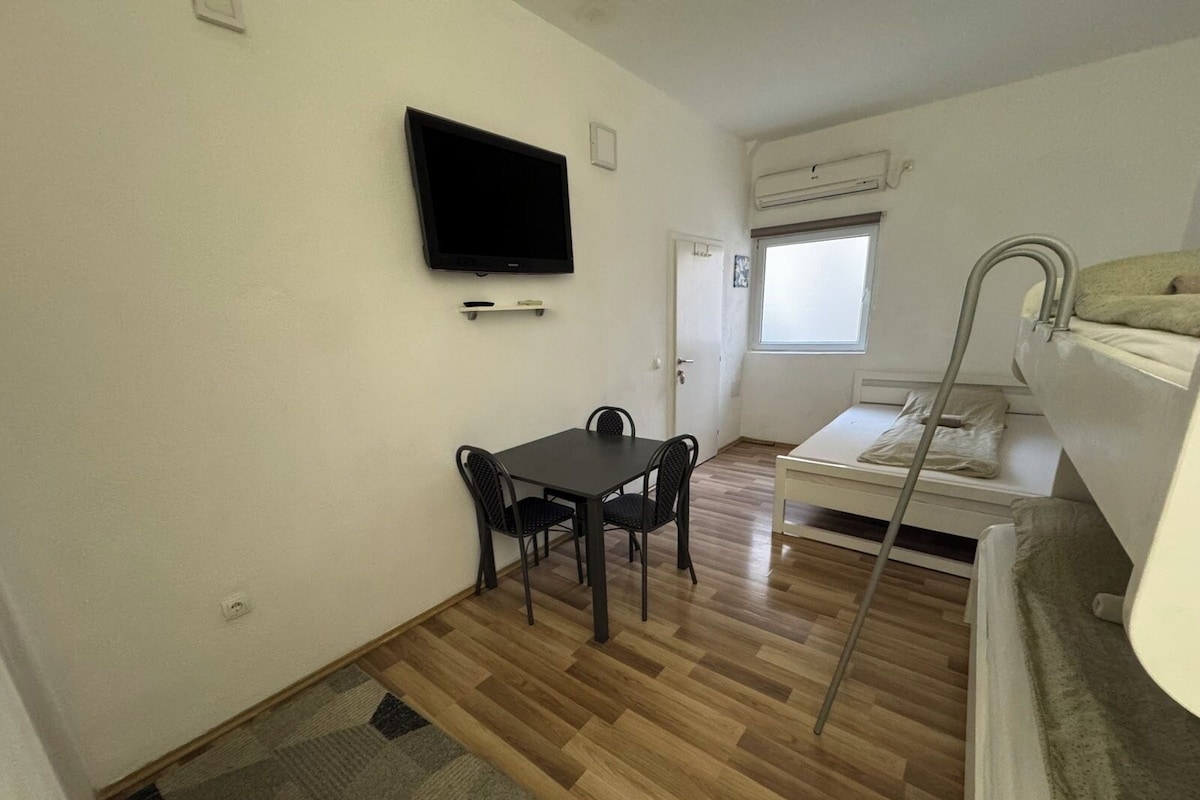 A small, compact room with a wooden floor, a single table with two chairs, a bed with a mattress and pillows, a TV mounted on the wall, and a window with a view.