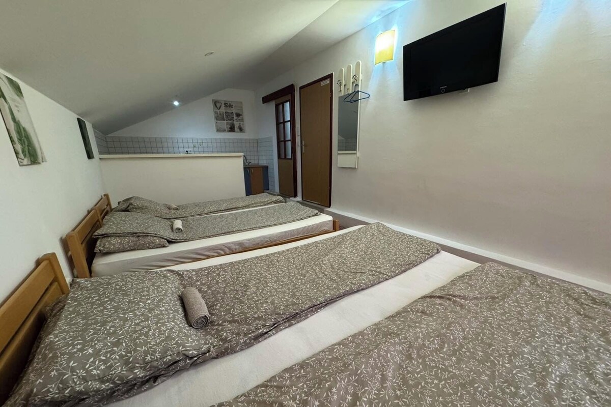 A room with three beds arranged in a row, a television mounted on the wall, and a small kitchenette area in the background.