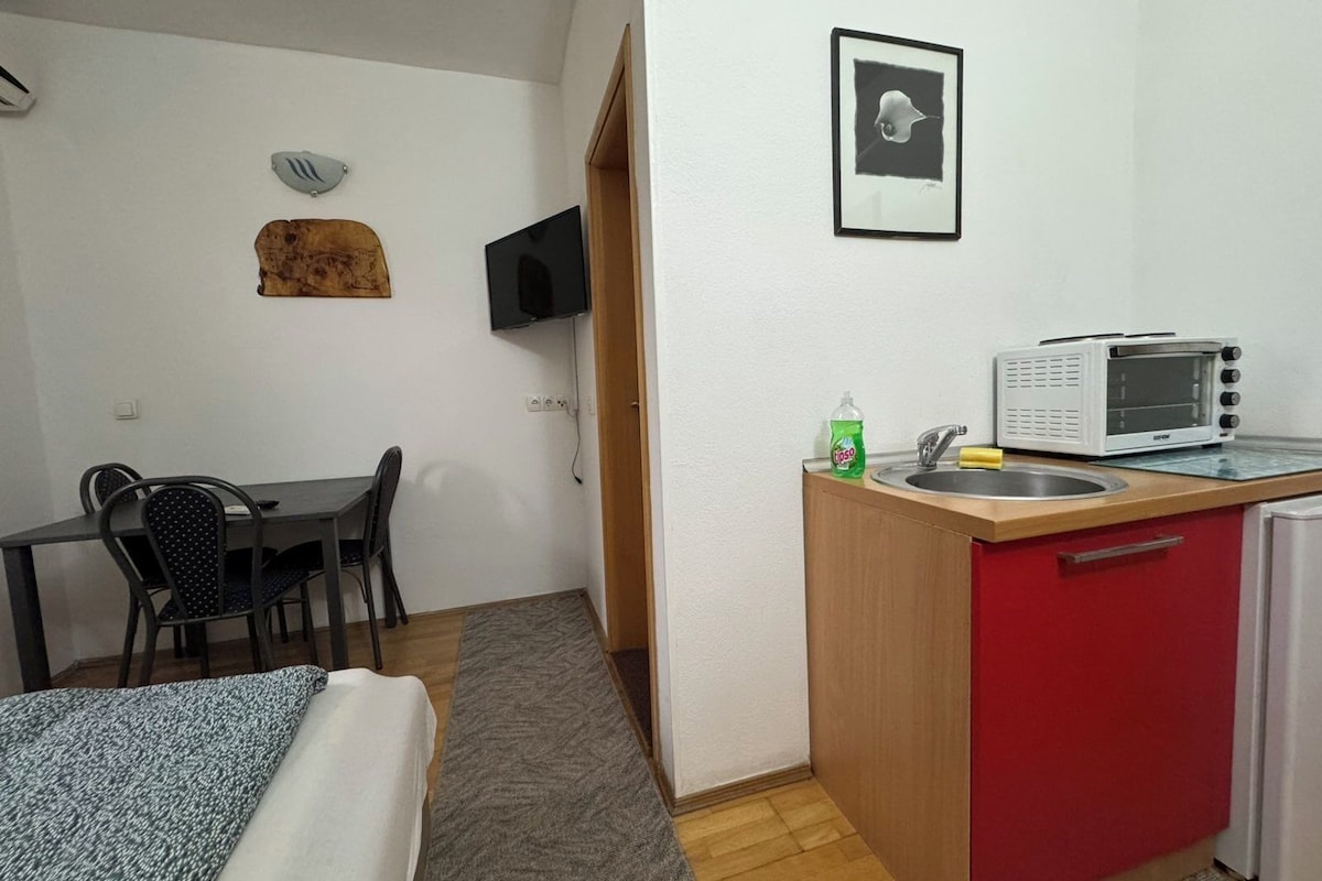 A compact room with a bed, a desk, a chair, a small kitchenette with a microwave, a sink, and a red cabinet.