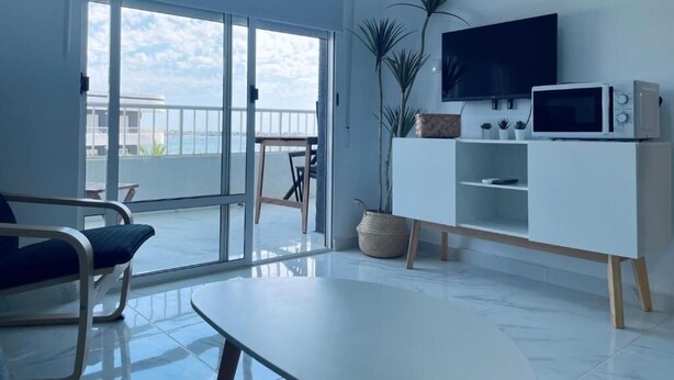 A modern living room with a large sliding glass door leading to a balcony, a flat-screen TV mounted on the wall, a white entertainment unit, and a round table. - Palmera Beach by Renthas (Orihuela)