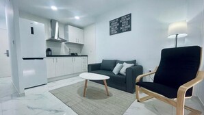 A modern living room with a sofa, a coffee table, and a chair. There is a kitchen area with cabinets and a refrigerator. A sign with text is visible on the wall. - Palmera Beach by Renthas (Orihuela)