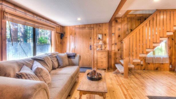 Interior acogedor de una cabaña con escalera de madera, un sofá, una pequeña mesa y un ventanal con vista a árboles.