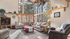 A cozy living room with two sofas, a coffee table, and a TV. Large windows offer a view of trees.