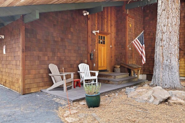 Una caba ña de madera con un porche, dos sillas Adirondack blancas, una planta en maceta, una bandera y un árbol.