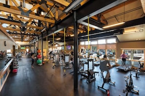 A spacious gym with various exercise equipment, large windows, and a high ceiling with exposed beams.