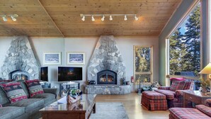 Un salón acogedor con chimenea de piedra, asientos cómodos, un televisor de pantalla plana y ventanas amplias con vistas a árboles.