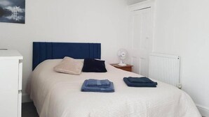 Quarto bem arrumado com cama, travesseiros, cobertores dobrados, mesa de cabeceira com abajur e radiador.