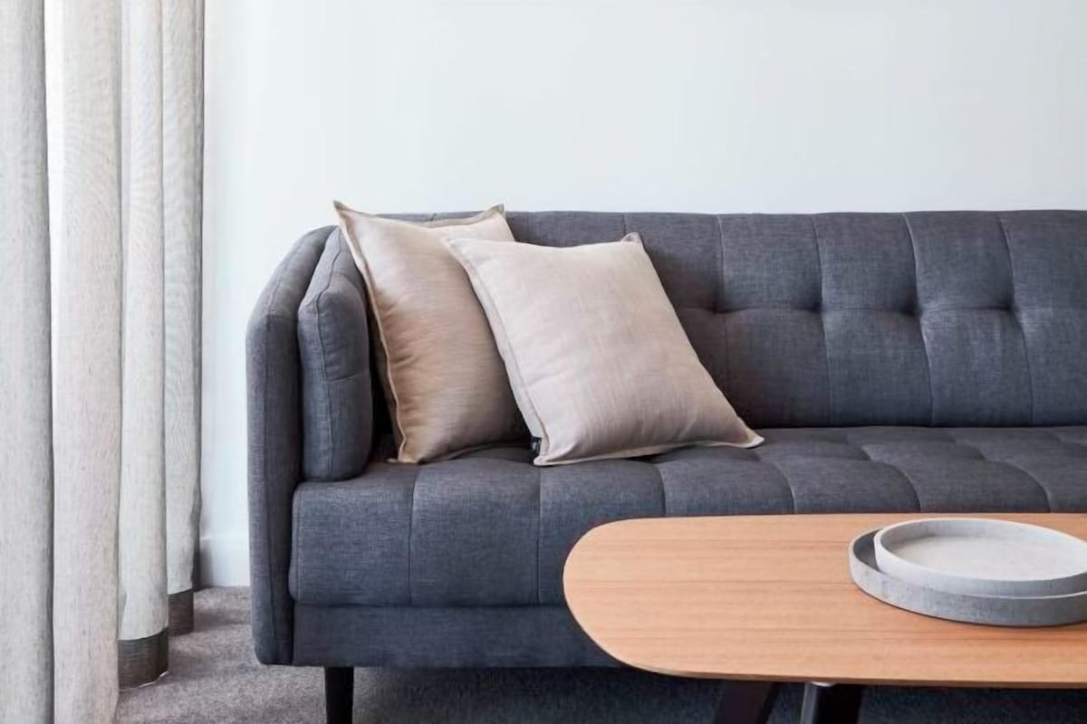 A grey sofa with two beige pillows, a wooden side table with a round tray, and a white curtain.