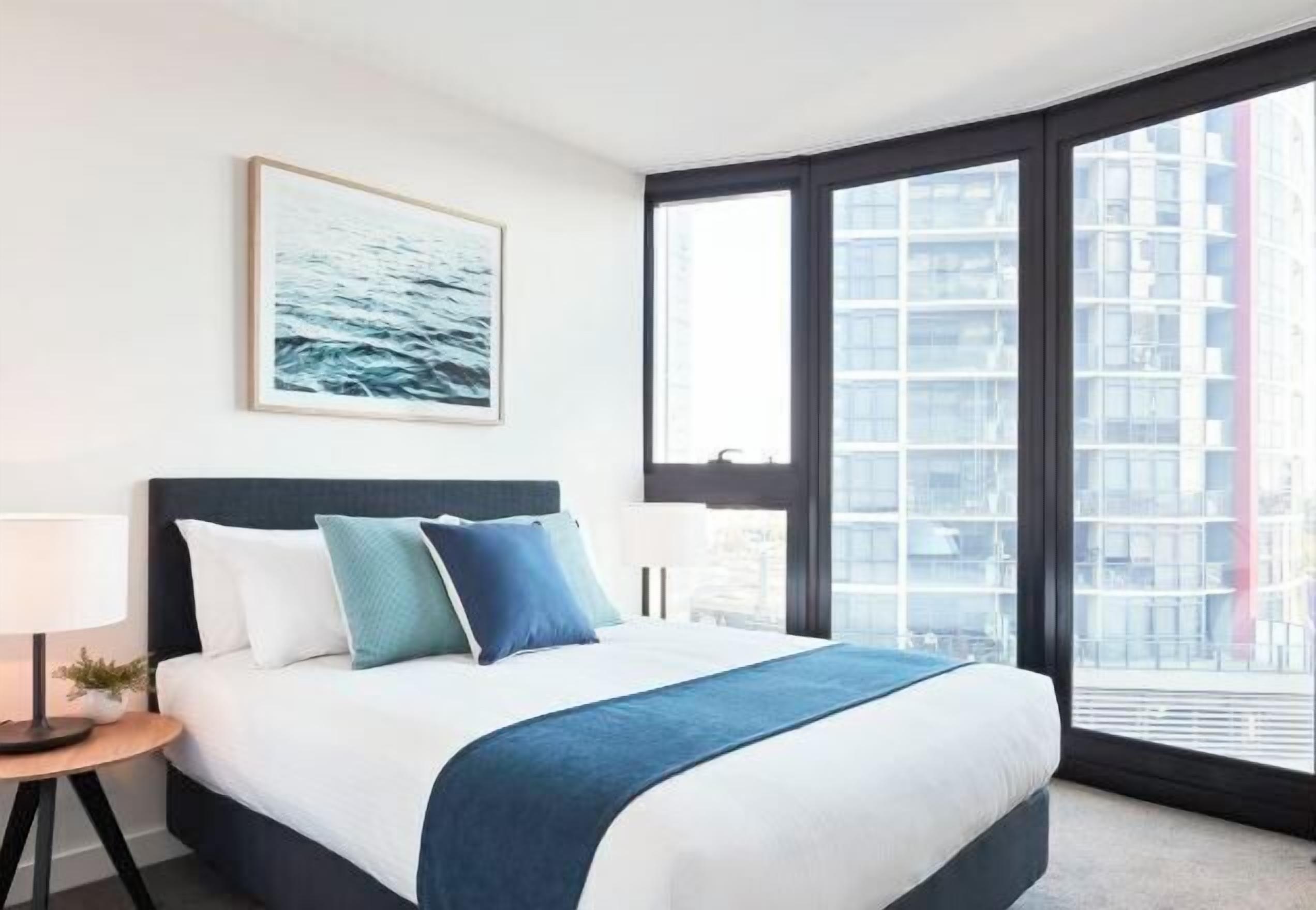Habitación de hotel con una cama grande, una mesita de noche, una lámpara, un cuadro en la pared y un amplio ventanal con vistas a edificios.