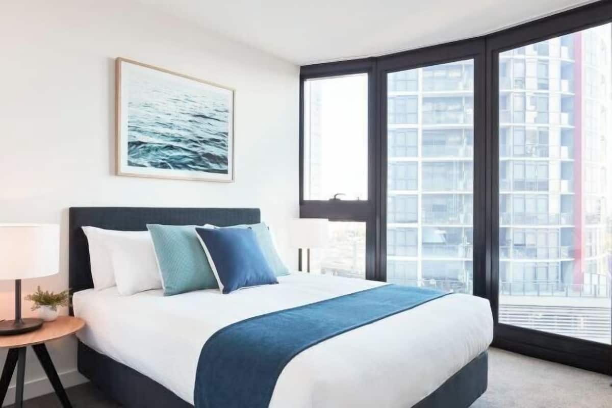 Une chambre d’hôtel avec un grand lit, une table de chevet, une lampe, un tableau encadré au mur et une grande fenêtre donnant sur des immeubles.
