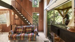 Un salon chaleureux avec un canapé à carreaux, une cheminée, un escalier en bois et de grandes fenêtres donnant sur l’extérieur.