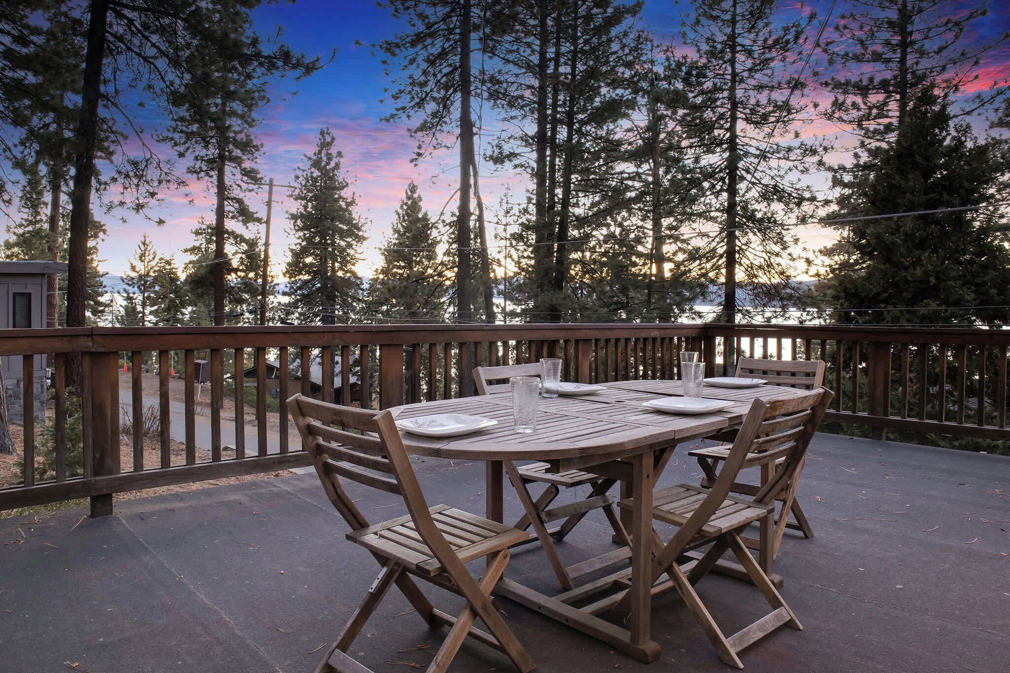 An outdoor wooden deck with a round table and chairs, surrounded by tall trees and a wooden railing.