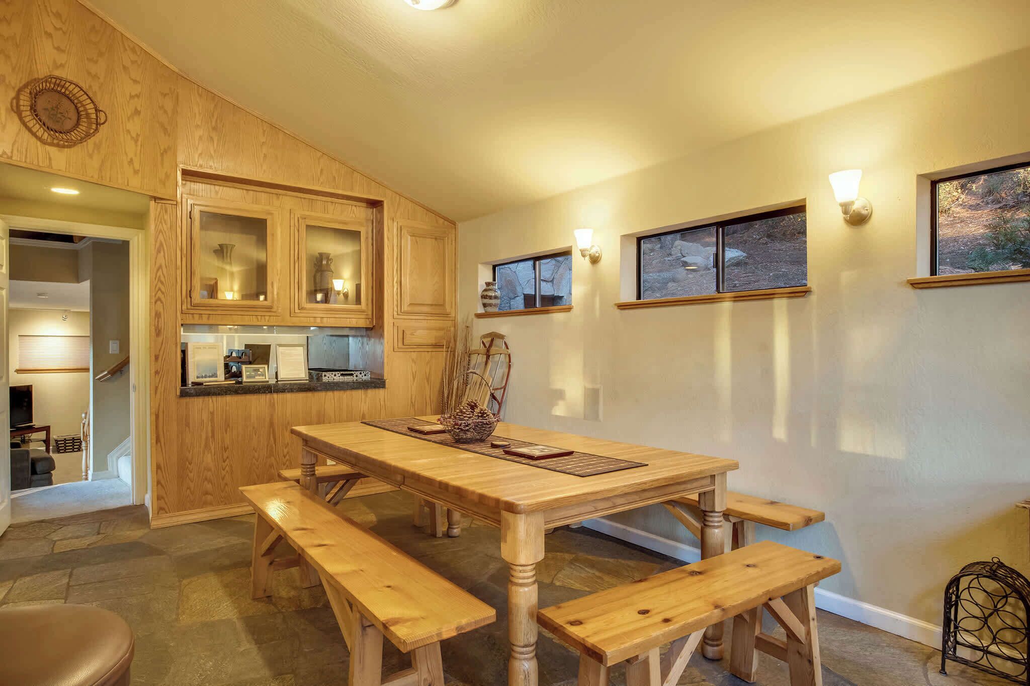 Une table à manger en bois avec des bancs, une cuisine avec des meubles en bois et une vue sur l’extérieur par les fenêtres.