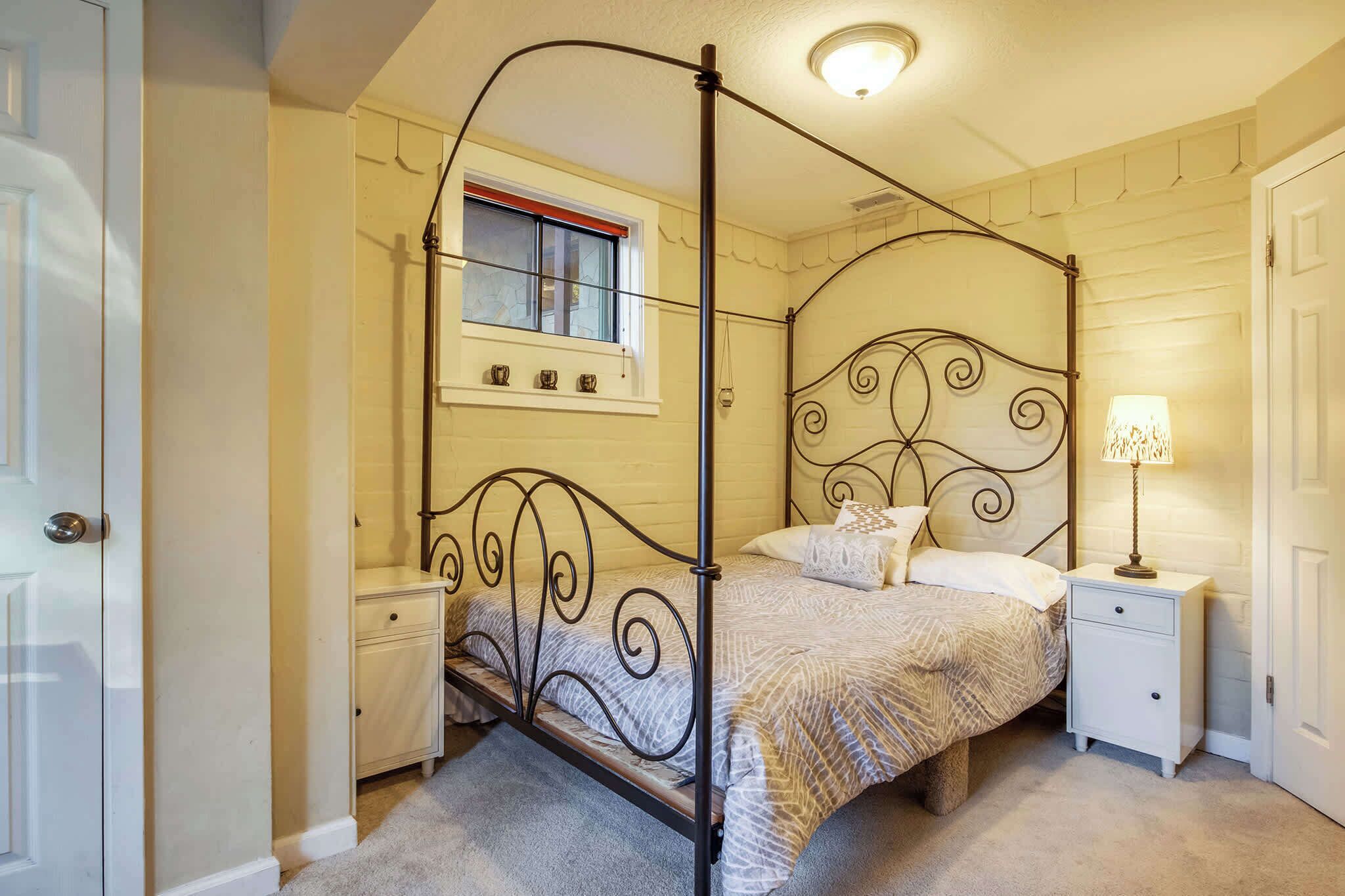 A four-poster bed with a metal frame, a bedside table, a nightstand, and a window with a red frame.