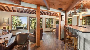 A spacious living area with a wooden ceiling, large windows, a sofa, a dining table, and a kitchen area with bar stools. - Snowshoe Cabin | 3 Decks, BBQ & Hot Tub! (Tahoe City)