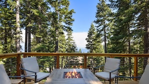 A deck with a fire pit table and four chairs, surrounded by tall trees. - Snowshoe Cabin | 3 Decks, BBQ & Hot Tub! (Tahoe City)