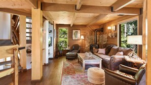 A cozy cabin interior with wooden beams, a fireplace, leather furniture, and a large window offering a view of the outdoors. - Snowshoe Cabin | 3 Decks, BBQ & Hot Tub! (Tahoe City)