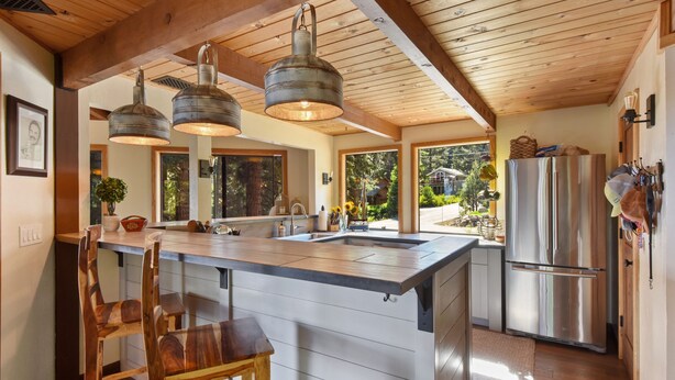 A kitchen with wooden beams, a large island with bar stools, a stainless steel refrigerator, and hanging pendant lights. - Snowshoe Cabin | 3 Decks, BBQ & Hot Tub! (Tahoe City)