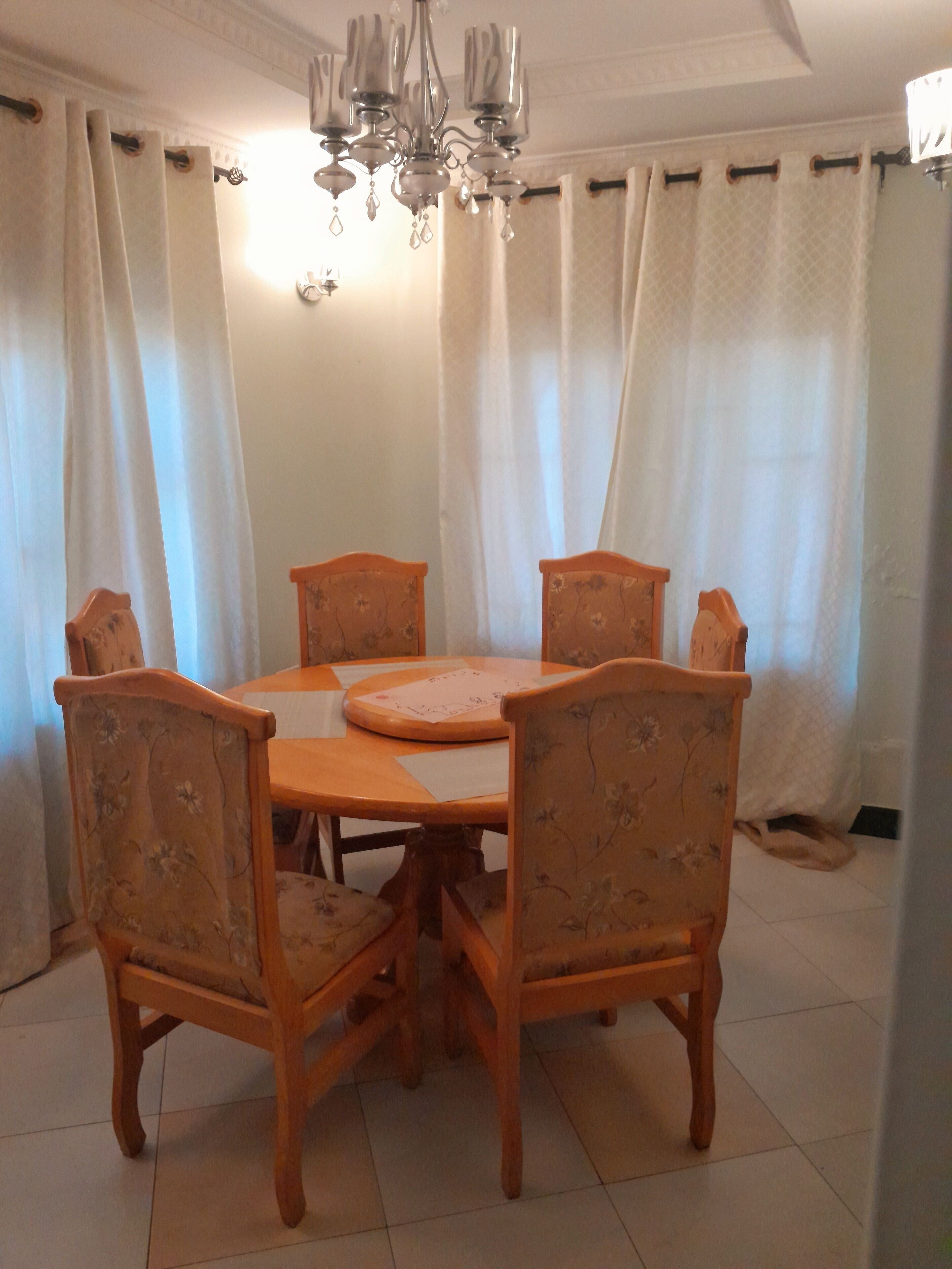 Comedor con una mesa redonda de madera y seis sillas, cada una con tapicería floral. Un candelabro cuelga del techo, y la habitación tiene cortinas blancas.