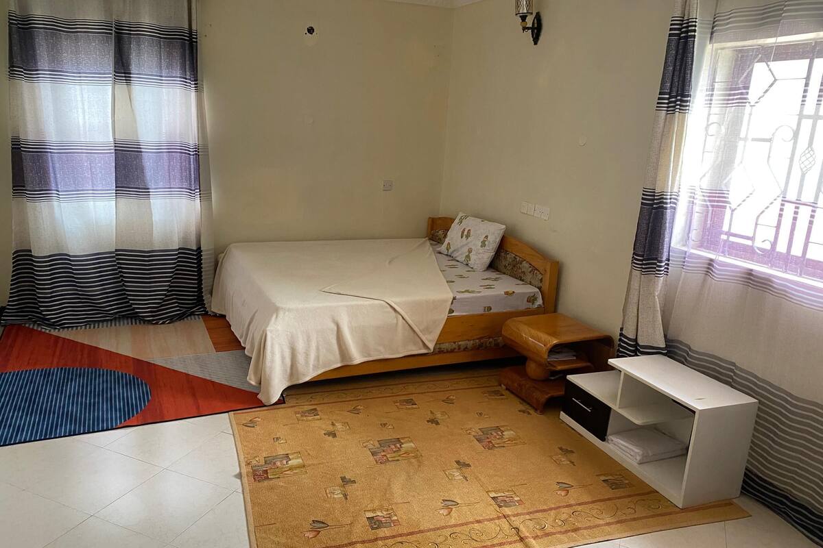 A small, neatly arranged bedroom with a single bed, a bedside table, a nightstand, a small shelf, and a window with curtains.