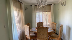 A dining area with a round table and six chairs, a chandelier, and a window with curtains. - anna's homestay (Arusha)