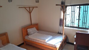 A single bed with a wooden frame and a mattress, a small bedside table, and a window with metal bars. - anna's homestay (Arusha)