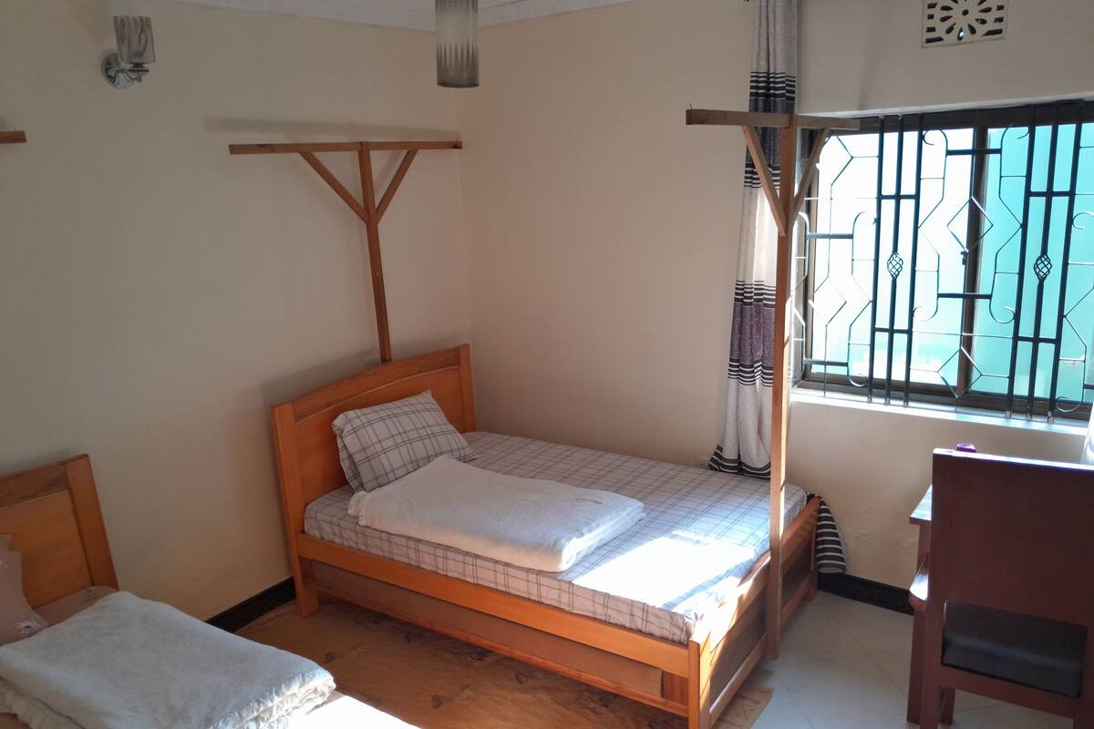 A single bed with a wooden frame and a mattress, a small bedside table, and a window with metal bars.