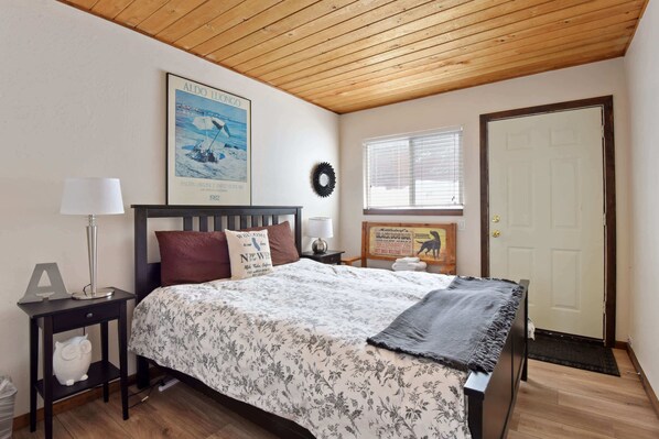 A bedroom with a wooden ceiling, a bed with floral bedding, a nightstand with a lamp, a framed welcome sign, and a door with a doormat.