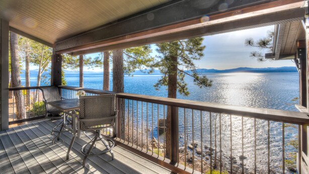 Eine Holzterrasse mit Tisch und Stühlen, mit Blick auf einen See, der von Bäumen und Bergen gesäumt wird.