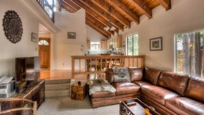 A spacious living room with a leather sofa, a wooden coffee table, and a TV. A kitchen is visible in the background with wooden cabinets and a dining area. - Glacier Forest Getaway | HOA Access! (Truckee)