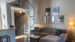 A living room with a curved staircase, a grey sofa, a coffee table, and a lamp. There are framed pictures on the wall and a sign above the staircase.