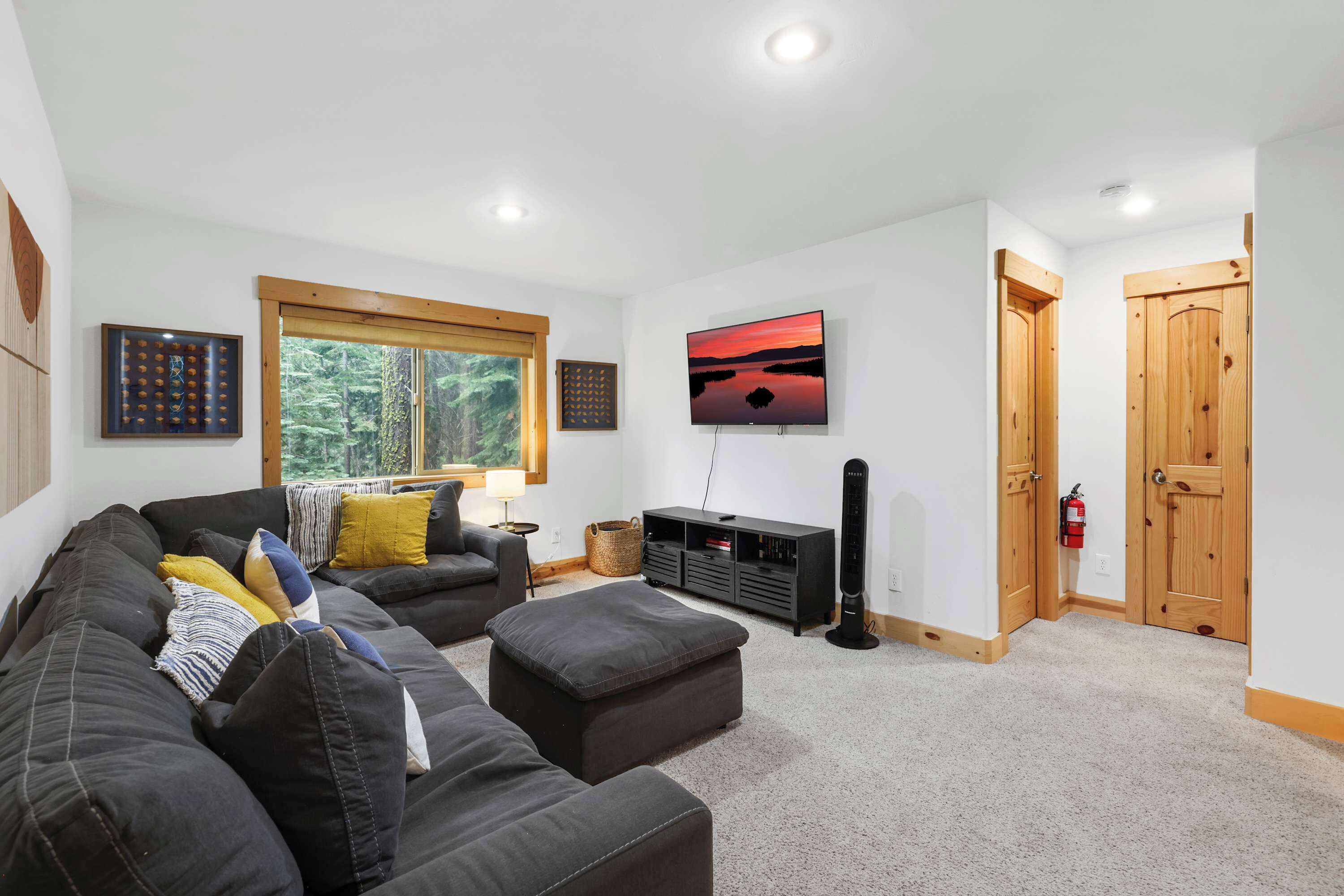 A modern living room with a large sofa, ottoman, and a flat-screen TV mounted on the wall. There is a window with a view of trees, a wooden door, and a fire extinguisher on the wall.