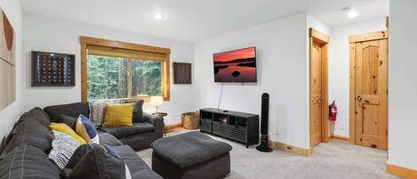 A modern living room with a large sofa, ottoman, and a flat-screen TV mounted on the wall. There is a window with a view of trees, a wooden door, and a fire extinguisher on the wall.