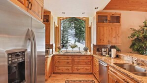 A kitchen with wooden cabinets, a stainless steel refrigerator, a built-in water dispenser, a granite countertop, a sink, and a window with a snowy mountain view.