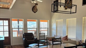 A spacious living area with a wooden dining table, chairs, and a ceiling fan. There are large windows with blinds, a fireplace, and a mounted deer head. - Home Sweet Home (Sevierville)