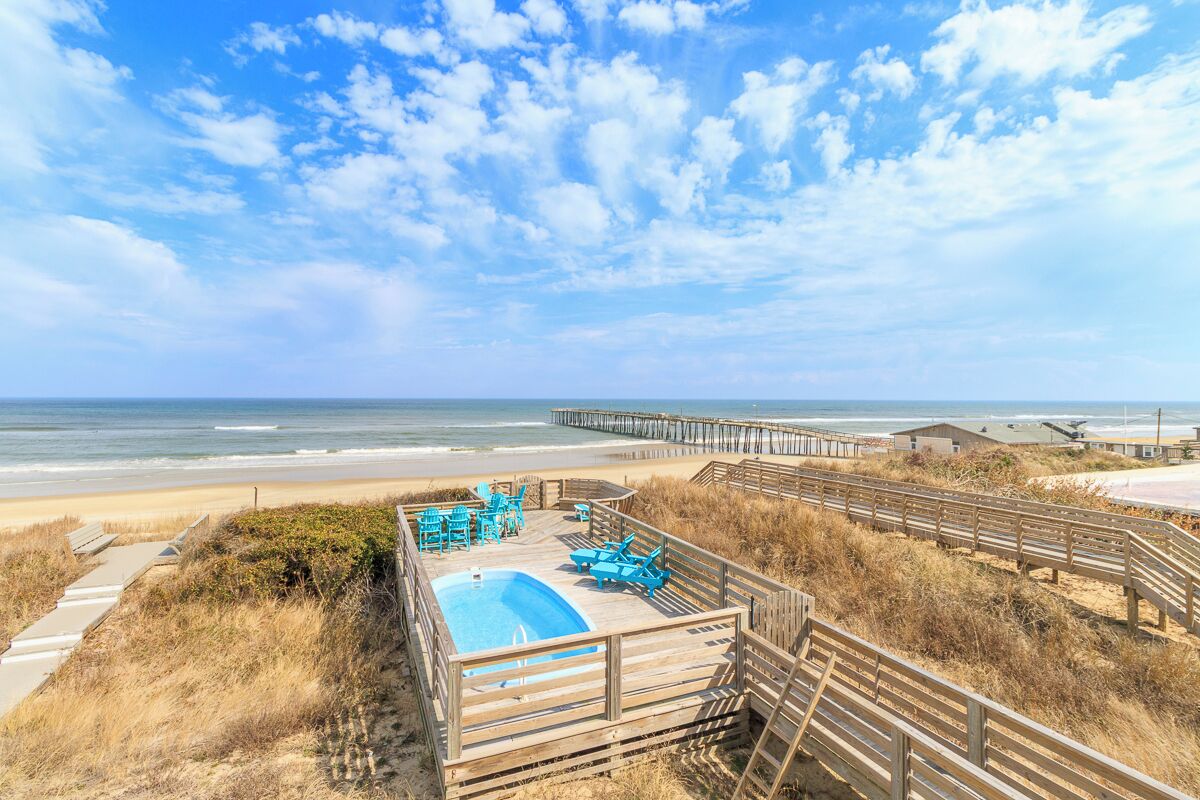 A beachside pool area with wooden deck chairs and a pool, overlooking the ocean.