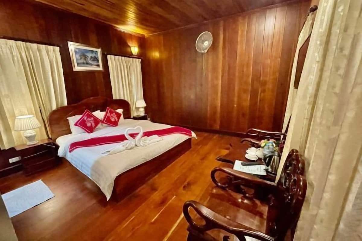 A wooden-paneled room with a bed, a desk, a chair, a fan, and curtains.