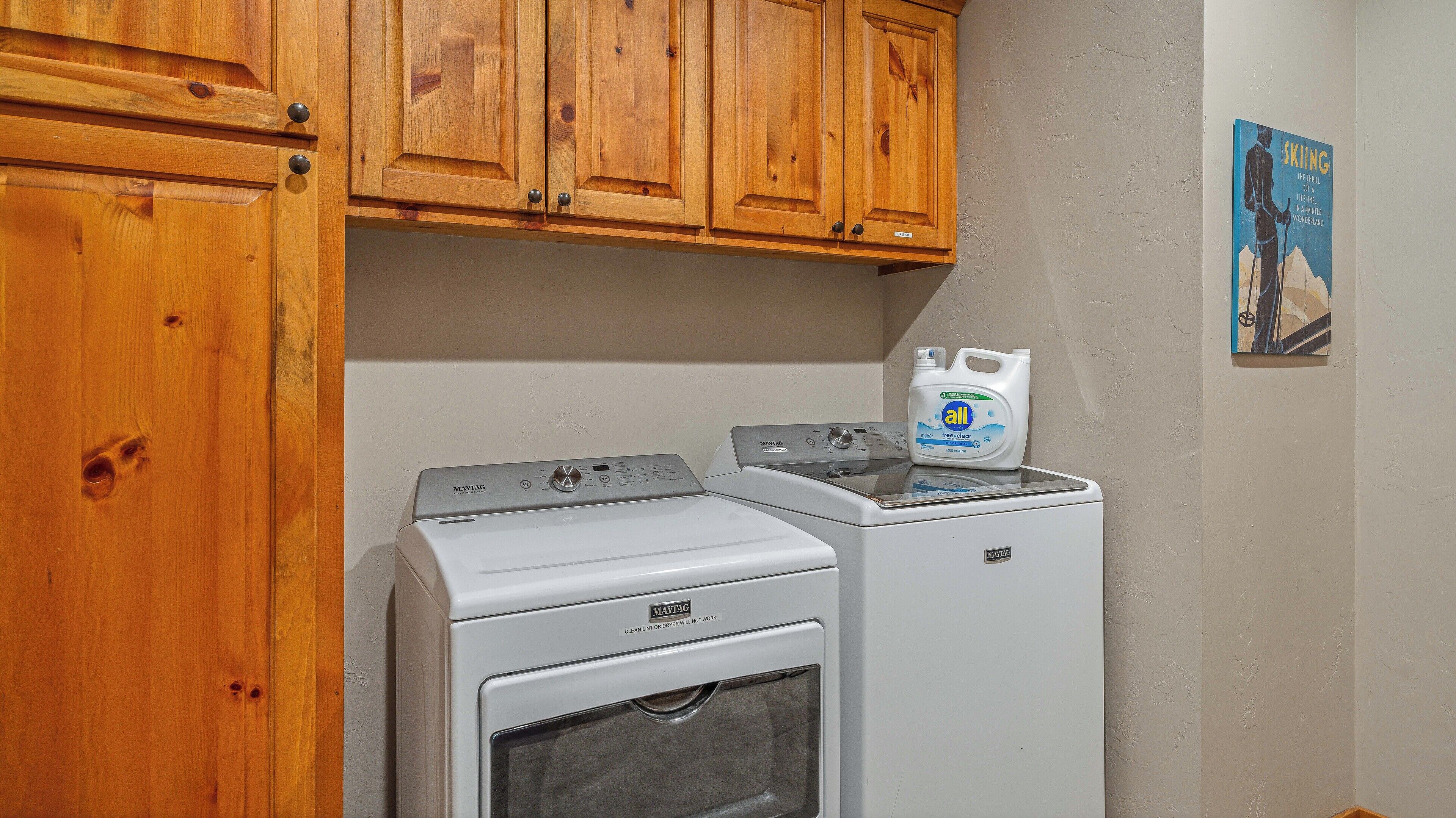A laundry area with two Maytag washing machines and a dryer, wooden cabinets above, and a poster on the wall.