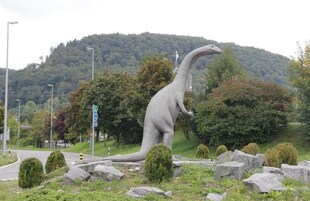 Una estatua de dinosaurio en un parque, con un camino y árboles al fondo.