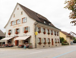 Un edificio de dos plantas con el nombre "Hotel Adler" en la fachada. Presenta un estilo arquitectónico europeo tradicional, con techo a dos aguas y ventanas de tejado. Hay zonas de estar exteriores con mesas y sillas frente al edificio.