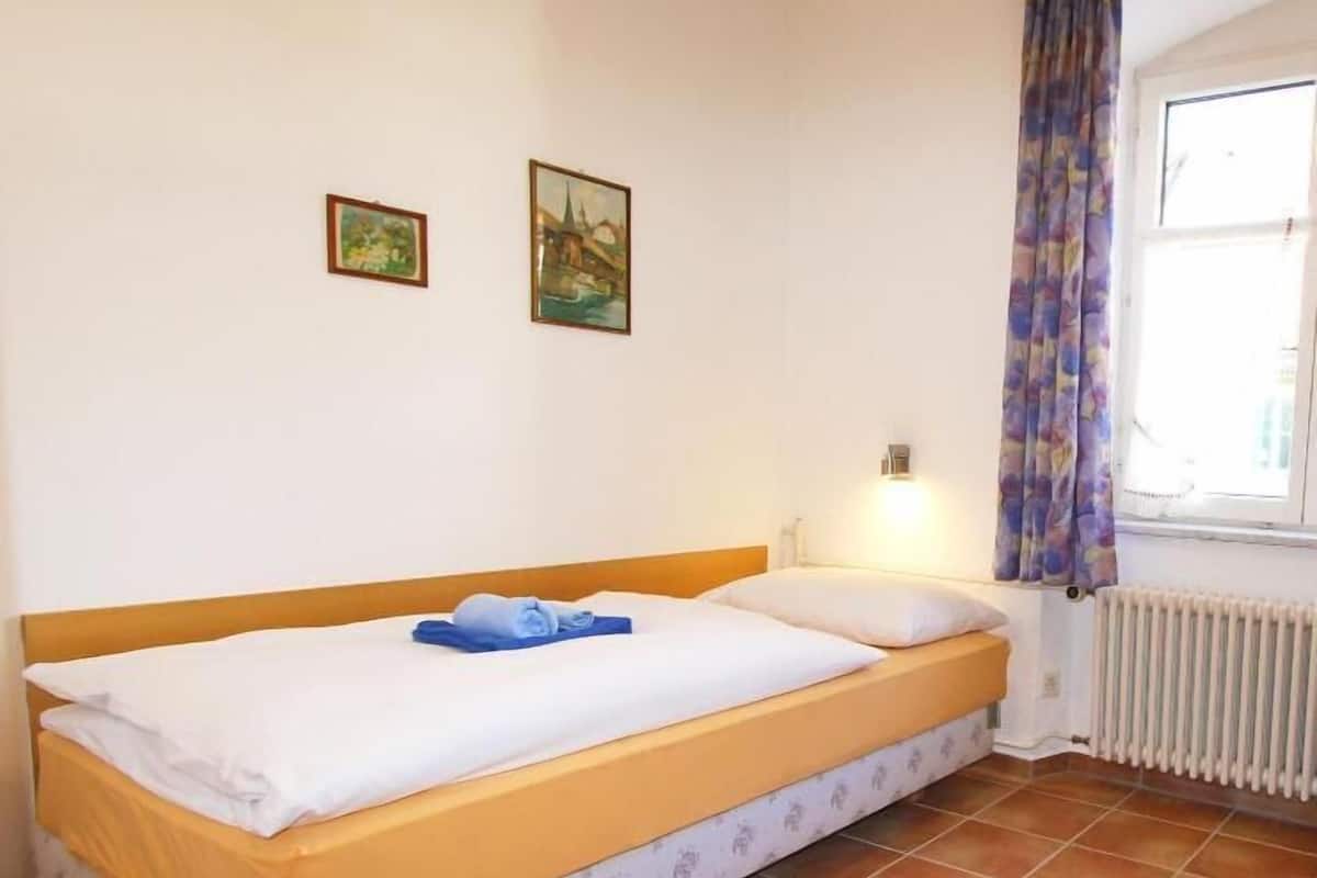 A single bed with a wooden headboard, a white mattress, and a blue towel. There are two framed pictures on the wall, a window with a curtain, and a radiator on the floor.