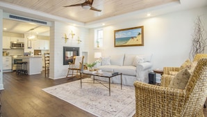 A spacious living room with a sofa, wicker chairs, a coffee table, and a painting on the wall. A kitchen with modern appliances is visible in the background. - Fabulous, 4 Bedroom, 3 Bathroom home in Downtown Ludington (Ludington)