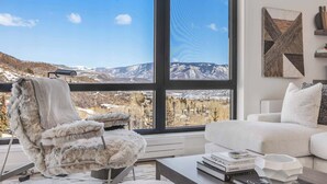 Een moderne woonkamer met een groot raam dat uitzicht geeft op bergen, een fauteuil, een witte bank en een salontafel met boeken.