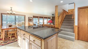 A kitchen with a granite countertop, wooden cabinets, a dining area with a table and chairs, a staircase with wooden railings, and large windows with a view of trees. - JACK01 - Family Friendly Sunriver Getaway (Sunriver)