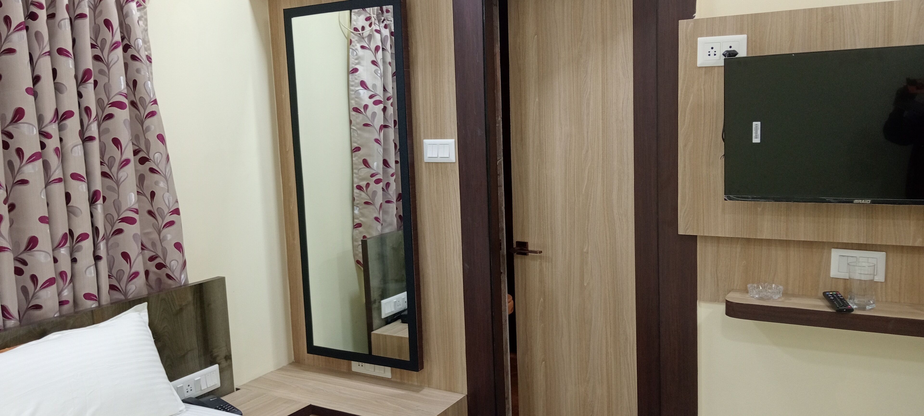 Habitación de hotel con cama, espejo, televisión y repisa con vasos y control remoto.