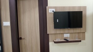 A room with a mounted flat-screen TV, a shelf with a remote control, and a door with a number sign. - Asha Inn (Barasat)