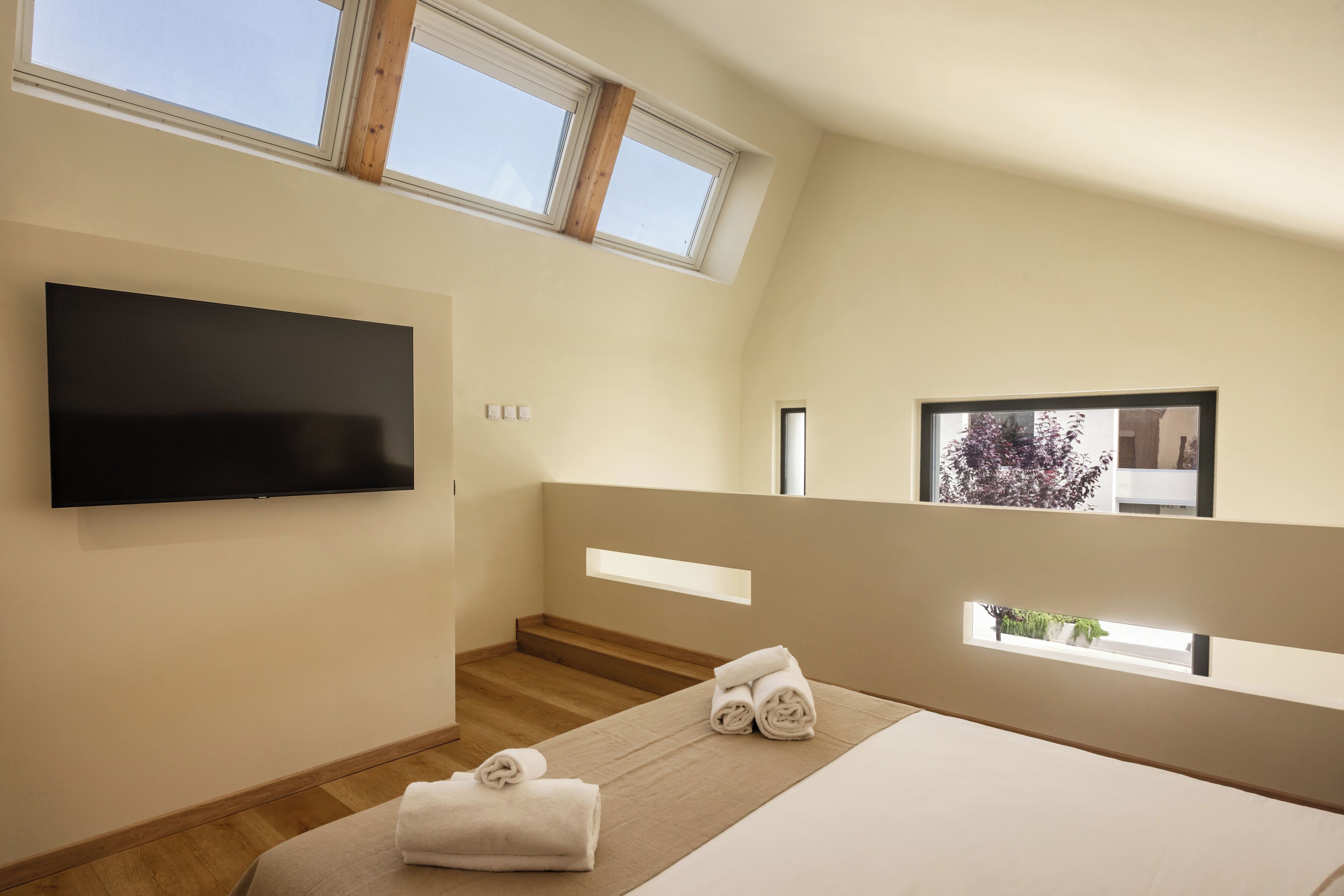 Une chambre moderne dotée d’un téléviseur à écran plat, d’un lit avec une tête de lit beige, d’un escalier en bois et d’une vue sur les arbres par la fenêtre.