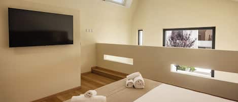 A modern bedroom with a flat-screen TV, a bed with a beige headboard, a wooden staircase, and a view of trees through a window.