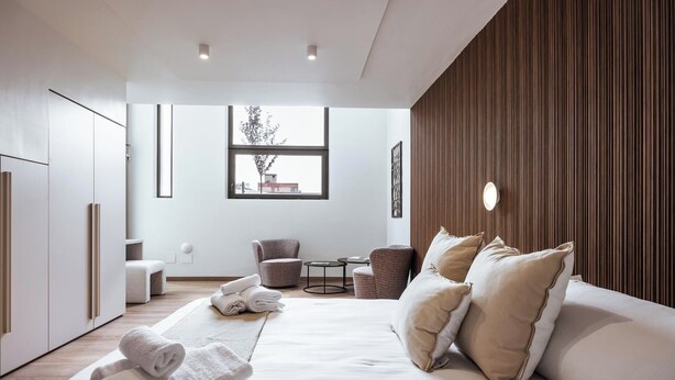 Een moderne hotelkamer met een groot bed, een zithoek met stoelen, een kleine tafel en een raam met uitzicht op gebouwen.