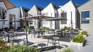Een terras buiten met meerdere tafels en stoelen, omringd door moderne gebouwen met grote ramen en een heldere lucht.